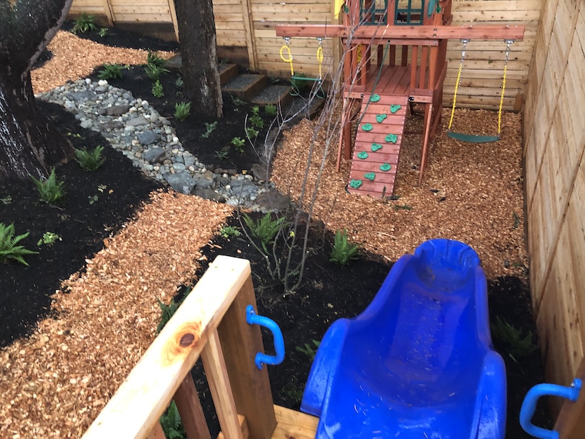 Sustainable backyard playground with wood mulch, rock path, and wooden playset with slide and swings