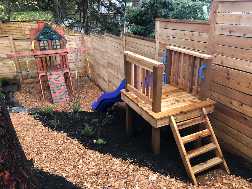 Wooden children's playset with slides and swings in landscaped backyard with mulch and plants
