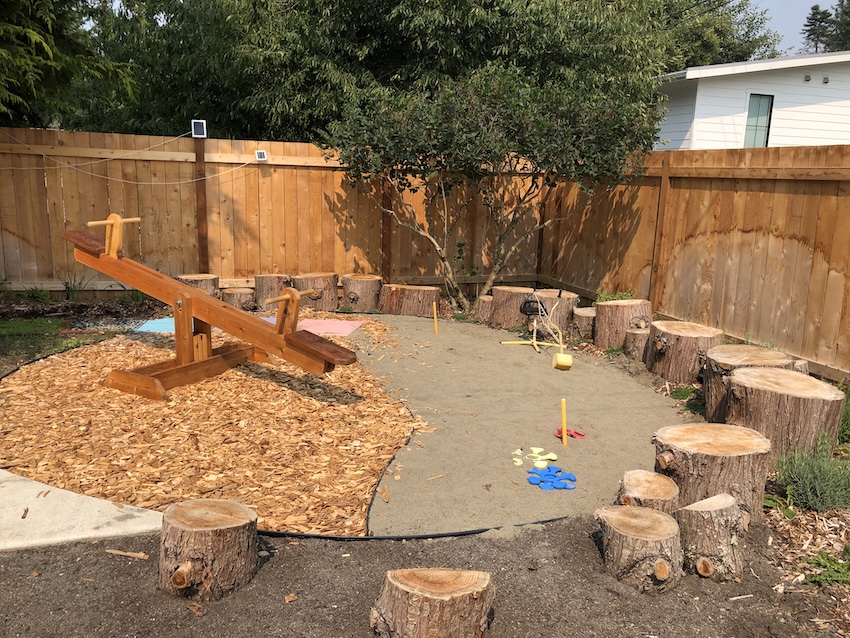 Sustainable backyard playground with wooden seesaw, mulch, sand, and tree stump seating area.