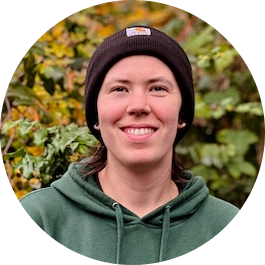 Smiling person in green hoodie and brown beanie with fall foliage background