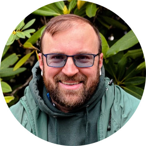 Smiling man with glasses in green jacket outdoors with leafy background